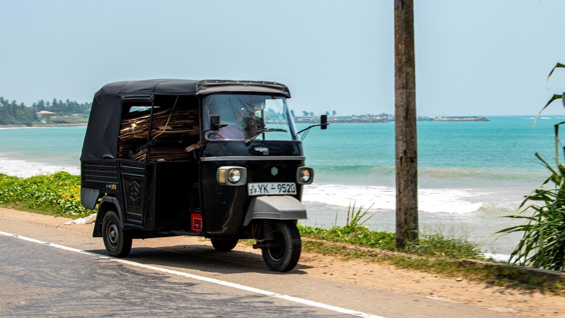 A black tuk-tuk parked on the coastal highway along Sri Lanka's south coast, with the Indian Ocean behind it